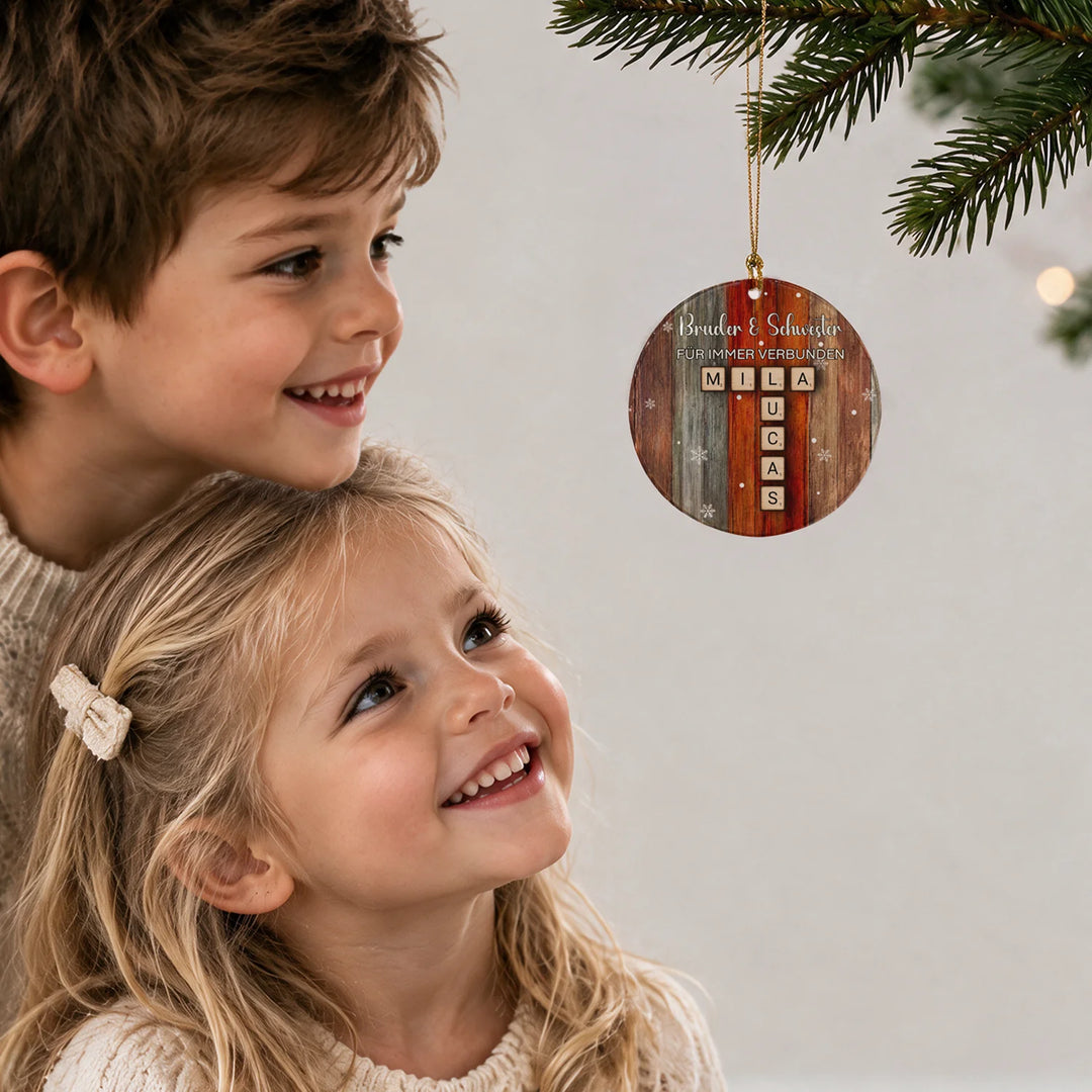 Bruder und Schwester schauen lächelnd auf den Scrabble-Anhänger „Bruder & Schwester“ am Weihnachtsbaum