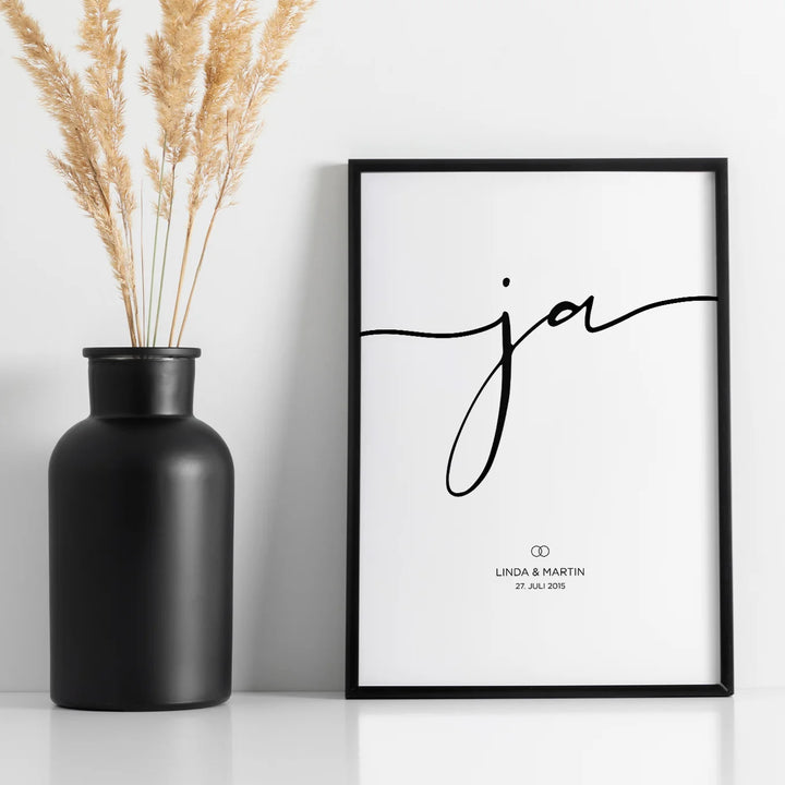 Personalisiertes Hochzeitsposter mit dem Wort "Ja" in eleganter Schrift, schwarzem Rahmen und Pampasgras-Dekoration in schwarzer Vase. Minimalistisches Design für ein stilvolles Hochzeitsgeschenk.